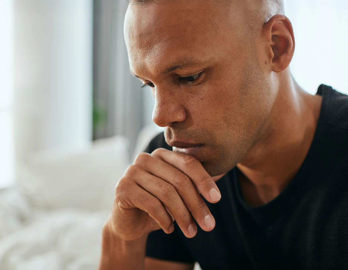 A close-up of a man in a black t-shirt looking downward with a troubled expression, resting his chin on his folded hand in a softly lit room.