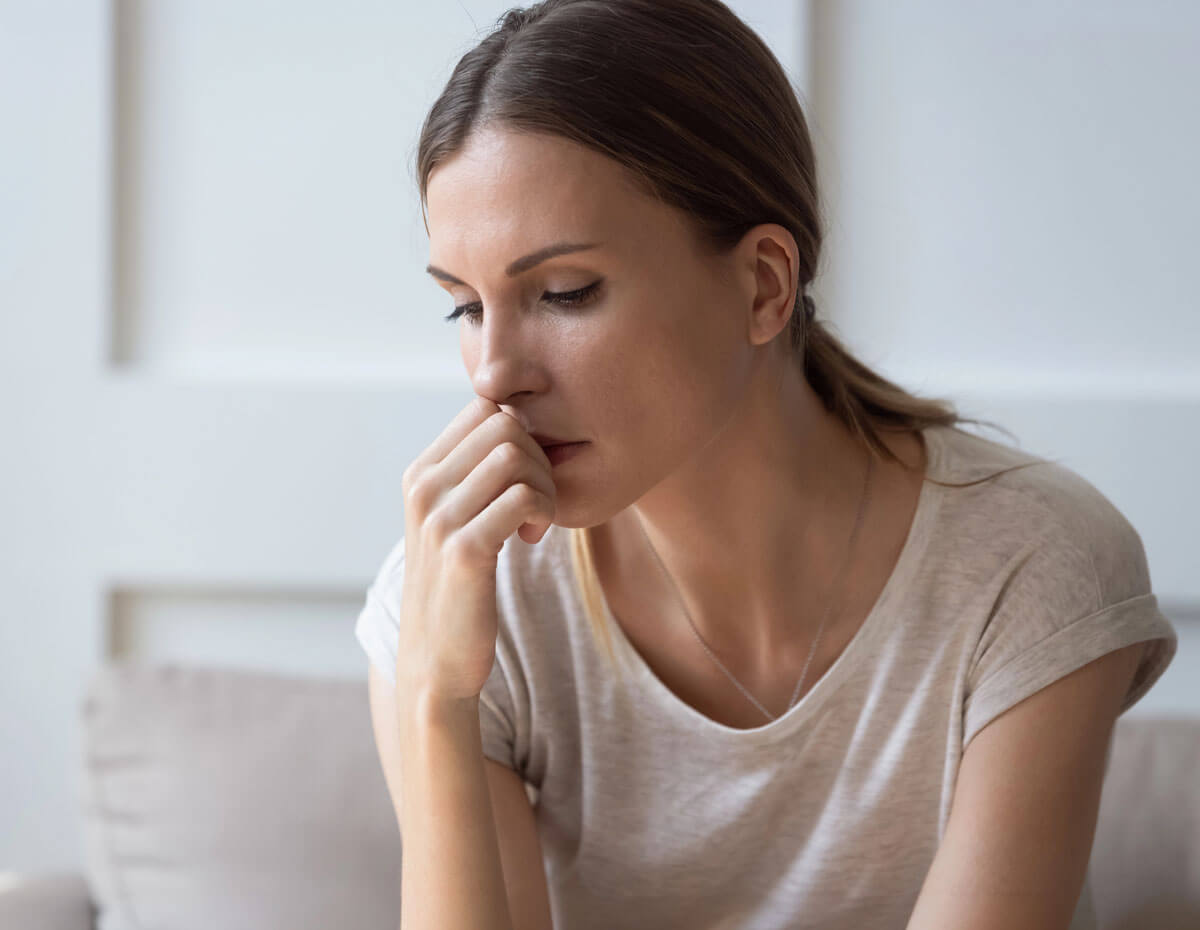 A young woman in a beige t-shirt sits with her eyes downcast and her hand raised to her face, conveying quiet worry or distress in a bright, neutral-toned room.